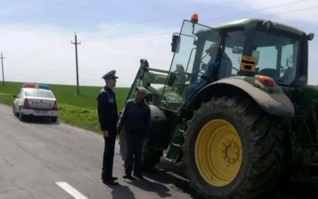 Un politist stă lângă un tractor verde cu roți mari galbene, lângă un alt individ care poartă o căciulă și o jachetă în fața unei mașini albe.