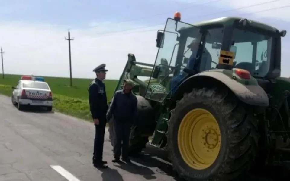 Un politist stă lângă un tractor verde cu roți mari galbene, lângă un alt individ care poartă o căciulă și o jachetă în fața unei mașini albe.