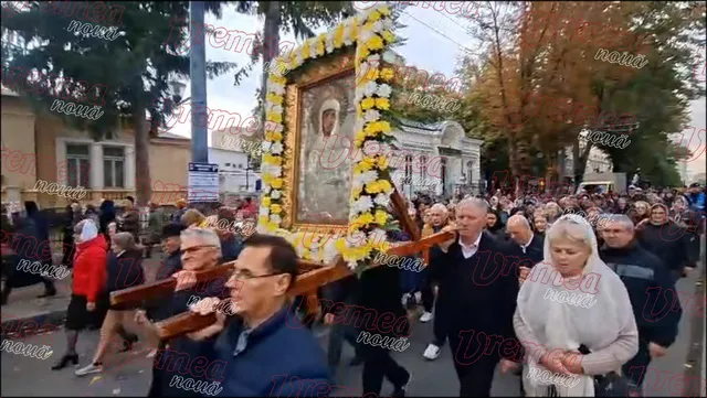 O mulțime de oameni merge pe o stradă lângă o icoană mare, decorată cu flori galbene și albe, purtată pe un suport de lemn.