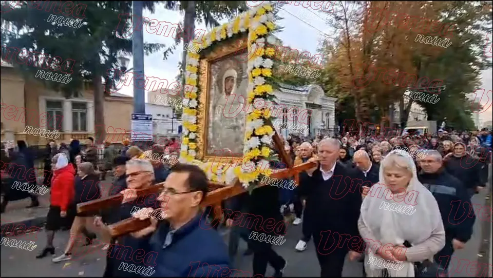 O mulțime de oameni merge pe o stradă lângă o icoană mare, decorată cu flori galbene și albe, purtată pe un suport de lemn.