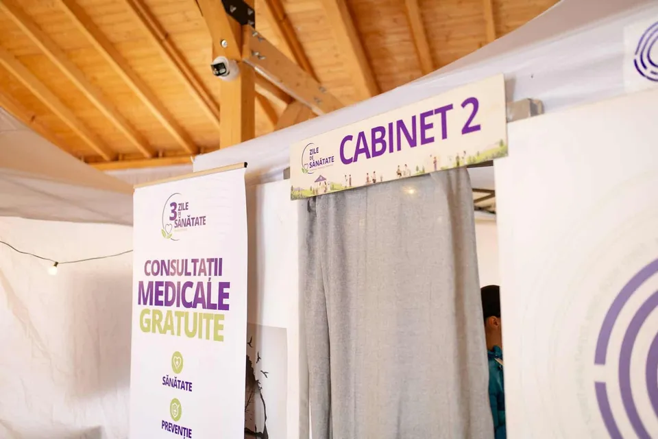 O cabină medicală etichetată „CABINET 2” cu un panou lateral care anunță consultații medicale gratuite, amenajată într-un spațiu cu acoperiș de lemn.