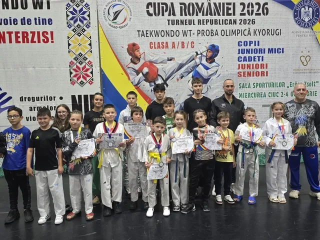 Un grup de copii și adolescenți stau în linie purtând medalii și diplome în fața unui panou publicitar colorat.