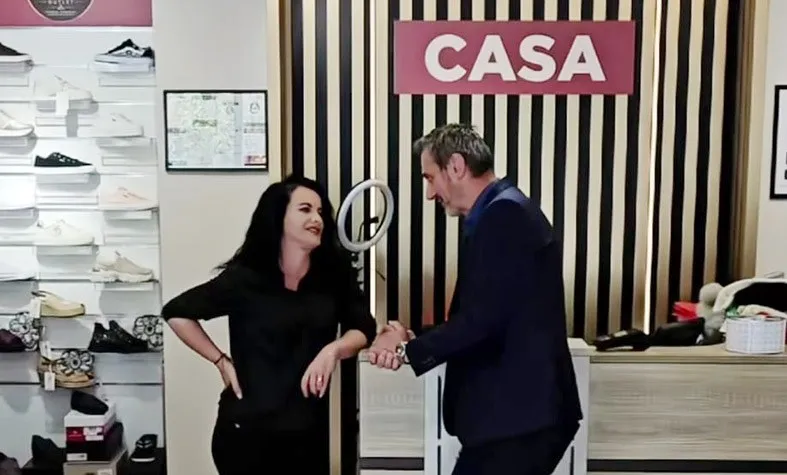 O femeie cu părul negru și un bărbat în costum întînd o mână într-un magazin cu rafturi de încălțăminte și un panou cu textul CASA.