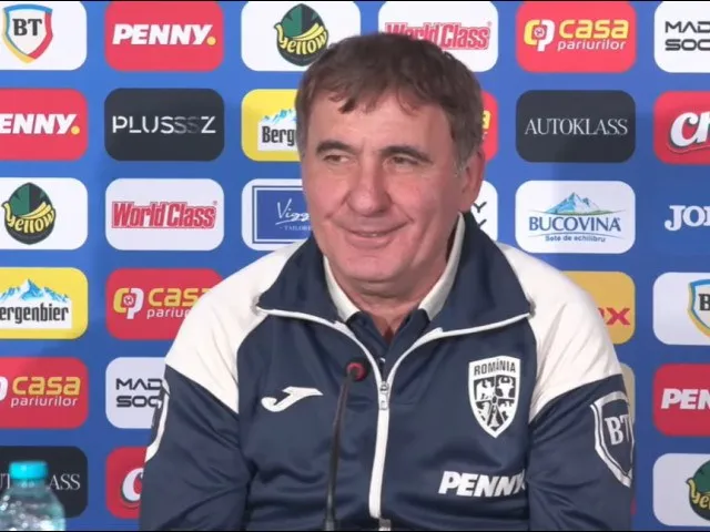 Un bărbat cu părul scurt și cămașă albastră sub un hanorac sportiv cu detalii albe stă în fața unui panou cu diverse logo-uri colorate.