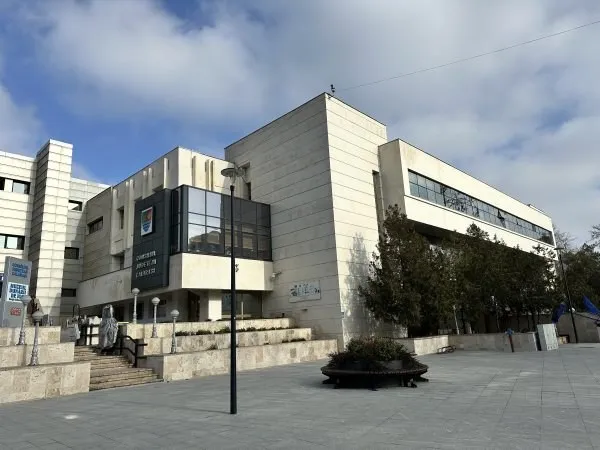 O clădire modernă din beton cu ferestre mari este situată lângă o zonă pietonală cu trepte și un stâlp de iluminat sub un cer înnorat.