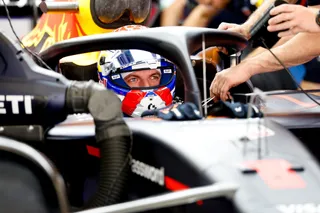 Un pilot de curse poartă o cască albastră și albă, fiind așezat în interiorul unui bolid de Formula 1.