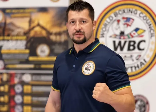 Un bărbat cu barbă poartă un tricou polo albastru cu embleme și stă cu un pumn închis ridicat în fața unui poster colorat.