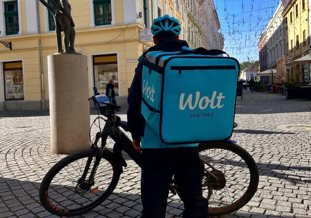 Un bărbat cu o cască albastră și un rucsac albastru cu inscripția Wolt stă lângă o bicicletă pe un drum pavat.