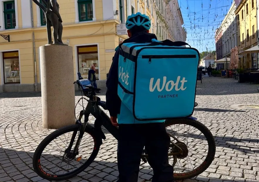 Un bărbat cu o cască albastră și un rucsac albastru cu inscripția Wolt stă lângă o bicicletă pe un drum pavat.