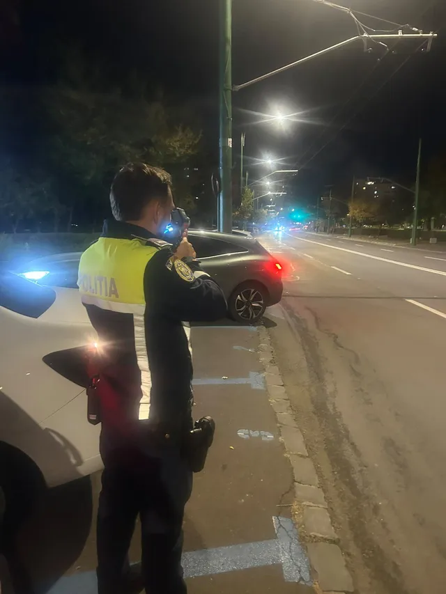 Un polițist cu vestă reflectorizantă stă lângă o mașină albă pe un drum urban iluminat de felinare noaptea.