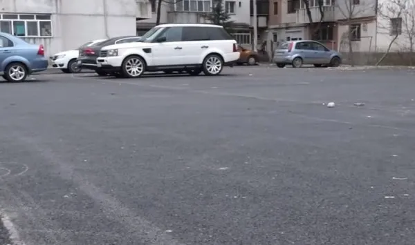 Mai multe mașini sunt parate pe un teren asfaltat lângă clădiri de apartamente.