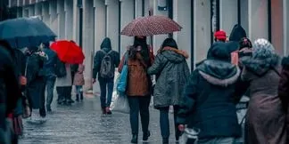 Oamenii merg pe un trotuar umed sub umbrele în timp ce poartă haine de iarnă și rucsacuri.