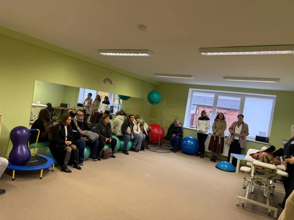 Un grup de persoane stă așezat pe scaune și pe mingi de fitness într-o încăpere cu pereți verzi și ferestre mari.