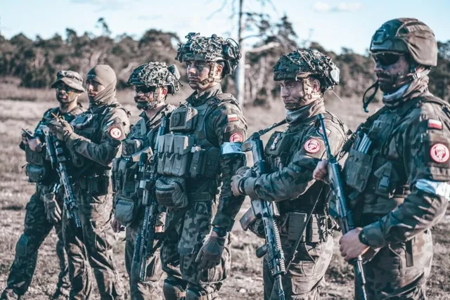 Șase soldați în uniforme de camuflaj și echipament militar stau în picioare pe un teren deschis sub un cer senin.