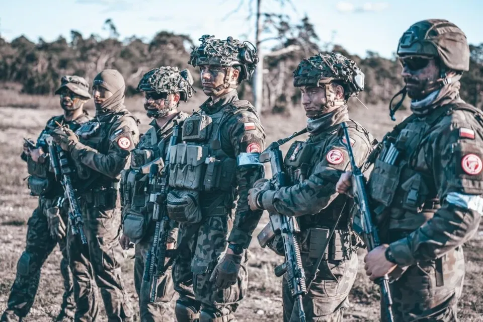 Șase soldați în uniforme de camuflaj și echipament militar stau în picioare pe un teren deschis sub un cer senin.