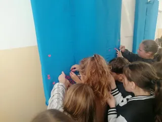 Un grup de copii stă în picioare și lipește mici forme roz pe un paravan de culoare albastră într-o încăpere interioară.