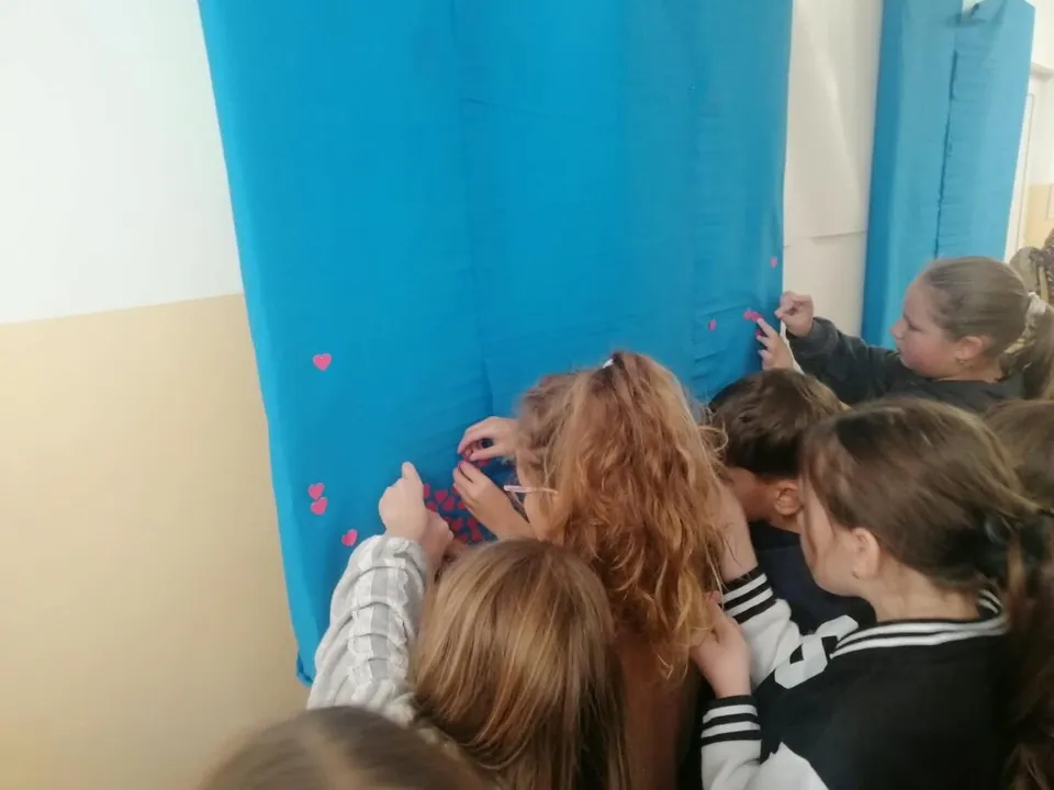 Un grup de copii stă în picioare și lipește mici forme roz pe un paravan de culoare albastră într-o încăpere interioară.