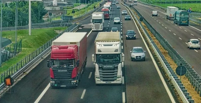 Mai multe camioane și mașini circulă pe o autostradă cu mai multe benzi de circulație, înconjurate de vegetație și bariere metalice de siguranță.