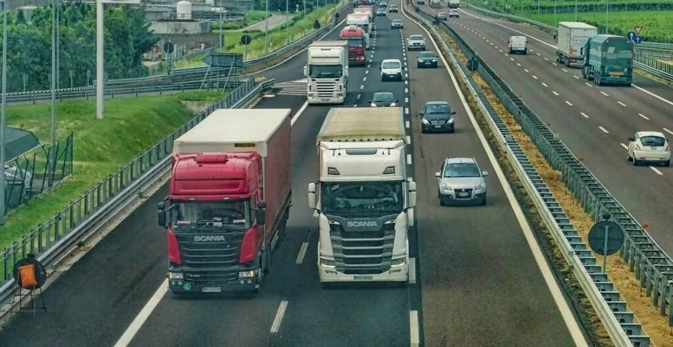 Mai multe camioane și mașini circulă pe o autostradă cu mai multe benzi de circulație, înconjurate de vegetație și bariere metalice de siguranță.