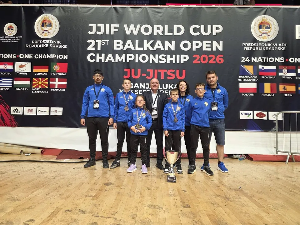 Un grup de sportivi în echipamente albastre stă în față pe un podium în fața unui panou negru cu inscripții albe și colorate.