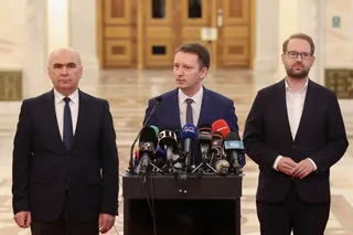 Trei bărbați în costume purtând cămași și cravate stau în spatele unui podium cu microfoane într-o încăpere cu uși de lemn.