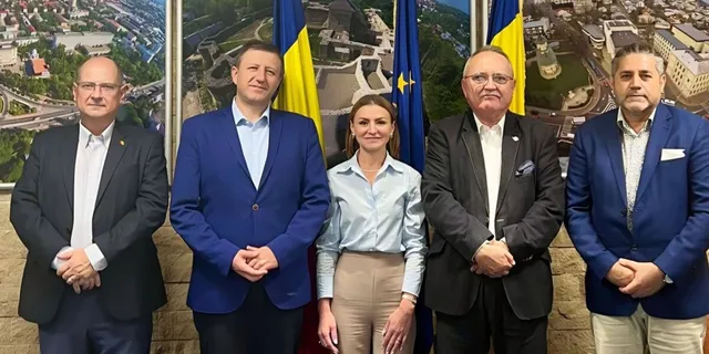 Cinci persoane stau aliniate în costume și ținute business, în fața unor drapele și panouri cu imagini urbane.