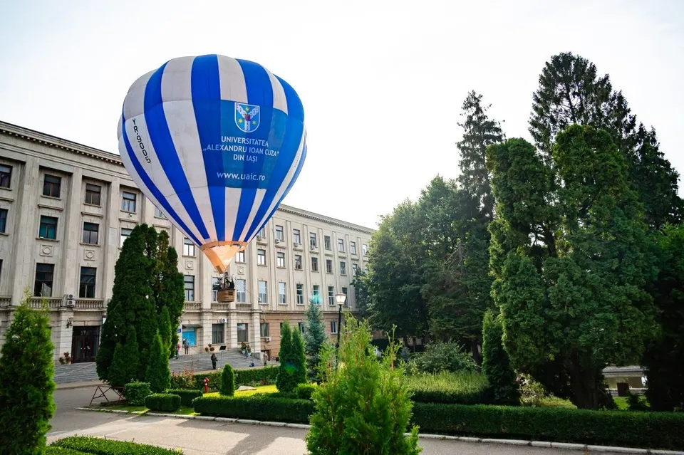 Un balon cu aer cald albastru și alb cu inscripții zboară deasupra unei clădiri mari de culoare deschisă și a unor copaci verzi.