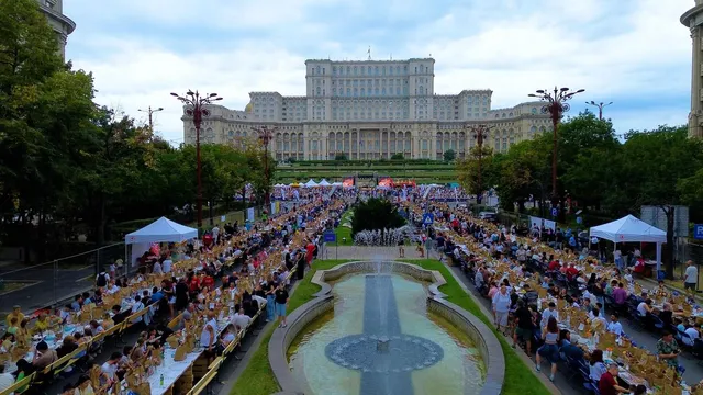 Oamenii stau la mese lungi așezate pe ambele părți ale unui fântâi, în fața unei clădiri mari și întinse sub un cer înnorat.