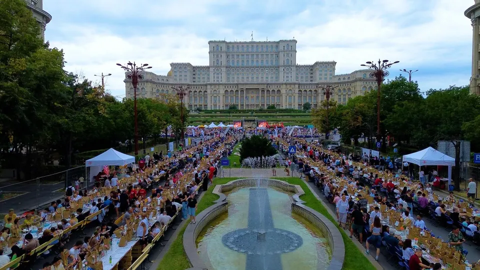 Oamenii stau la mese lungi așezate pe ambele părți ale unui fântâi, în fața unei clădiri mari și întinse sub un cer înnorat.