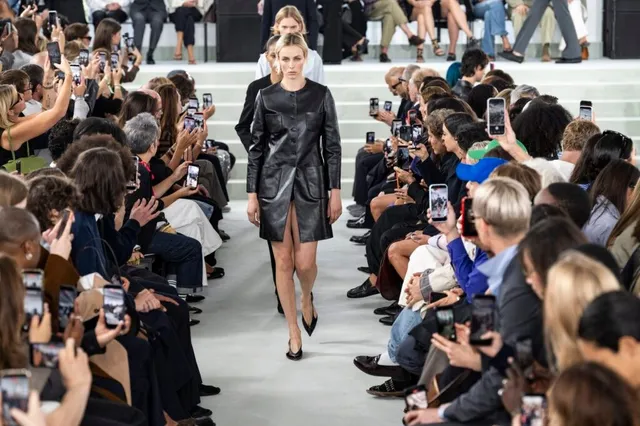 O femeie cu părul scurt și blond merge pe o pistă de catwalk purtând o rochie neagră din piele, înconjurată de spectatori care folosesc telefoane mobile.