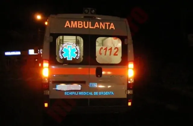Spatele unei ambulanțe albe cu inscripții portocalii și un simbol medical albastru este vizibil pe un fundal întunecat.