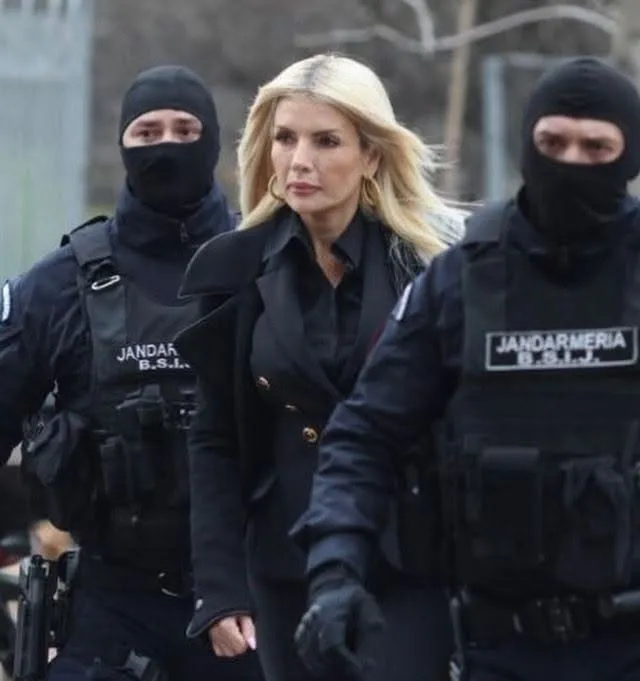 O femeie cu părul blond și haine negre merge între doi bărbați în uniformă neagră care poartă măști și echipament tactic.