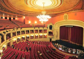 Interiorul unui teatru cu scaune roșii dispuse în curbe, un spectacol pe scenă și un candelabru mare suspendat de tavanul decorat.