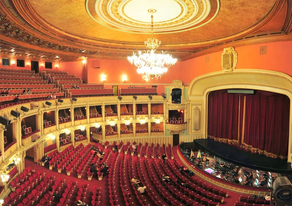 Interiorul unui teatru cu scaune roșii dispuse în curbe, un spectacol pe scenă și un candelabru mare suspendat de tavanul decorat.