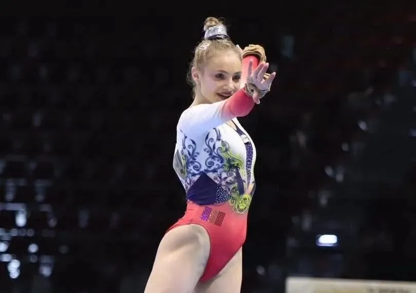 O gimnastă într-un costum de competição alb, albastru și roz stă în picioare cu mâinile ridicate într-o arenă întunecată.
