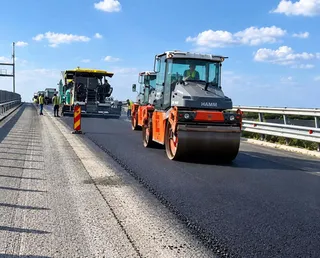 Un aparat de compactat de culoare gri și portocalie circulă pe o porțiune de asfalt proaspăt lângă un utilaj de pavaj sub un cer senin.