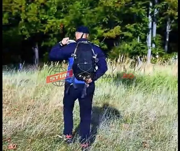 Un bărbat cu un rucsac negru și pantaloni închiși stă în picioare într-o zonă cu iarbă înaltă și copaci în fundal.