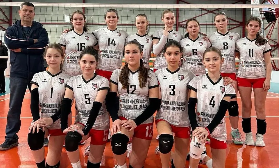 O echipă de sportivi în uniformă albă și roșie posedă pentru o fotografie de grup pe un teren de volei, alături de un bărbat.