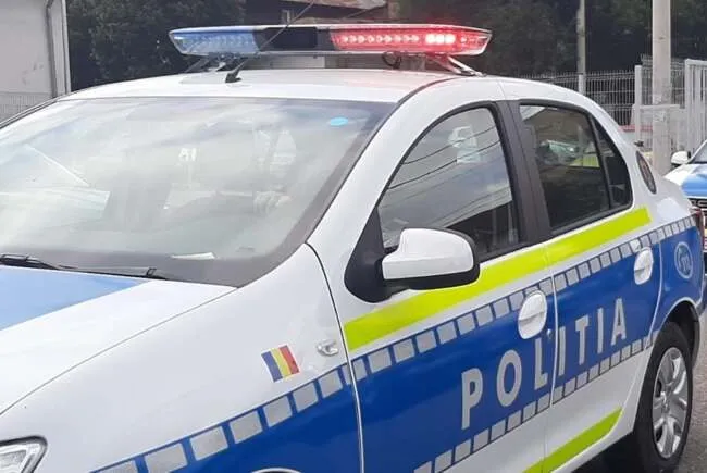 Un autovehicul alb cu marcaje albastre și galbene pe lateral, având inscripția POLITIA și girofaruri colorate pe plafon, este parcat în exterior.