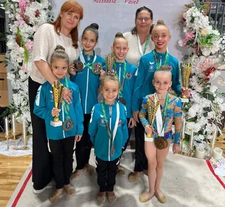 Șase gimnaste cu treninguri albastre și medii, alături de două femei, pozează cu trofee și medalii în fața unor aranjamente florale albe.