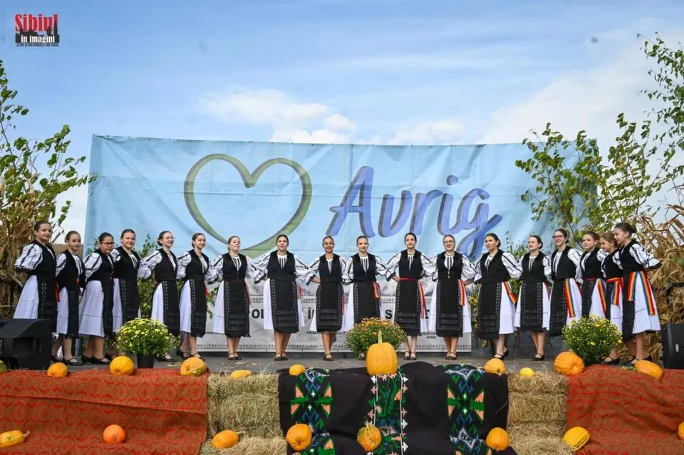 Un grup de femei îmbrăcate în costume populare albe cu detalii negre stau în rând pe o scenă decorată cu dovleci și plante verzi.