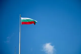 Un steag cu dungi orizontale alb, verde și roșu flutură pe un stâlp sub un cer albastru.