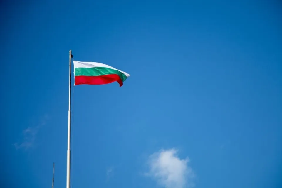 Un steag cu dungi orizontale alb, verde și roșu flutură pe un stâlp sub un cer albastru.