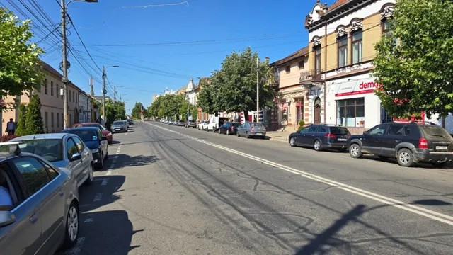 O stradă asfaltată cu linii albe este deschisă, având mașini parcate pe margini și clădiri cu ferestre mari pe partea dreaptă sub un cer senin.