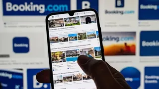 O persoană ține un smartphone care afișează o listă de anunțuri de cazare, în fața unui ecran mare cu diverse logouri Booking.com.