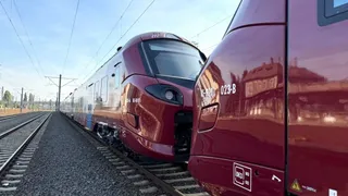 Un tren de culoare vișinie este oprit pe o cale ferată sub linii electrice aeriene, fiind filmat de lângă un alt vagon similar.