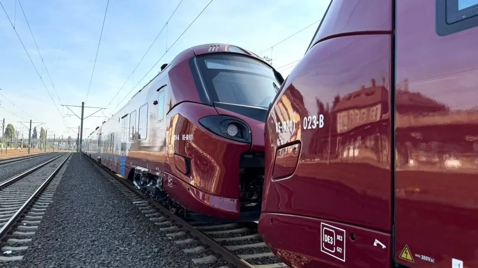Un tren de culoare vișinie este oprit pe o cale ferată sub linii electrice aeriene, fiind filmat de lângă un alt vagon similar.
