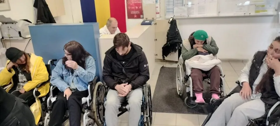 Cinci persoane stau în scaune cu rotile într-o încăpere cu pereți albi și diverse obiecte în fundal.