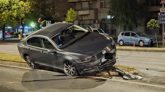 Un autoturism gri este poziționat pe un scuar de beton, având partea din față deteriorată și un obiect metalic de formă curbată în fața sa.