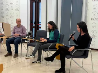 Trei persoane stau așezate pe scaune într-o încăpere, una dintre ele ținând un microfon în mână.
